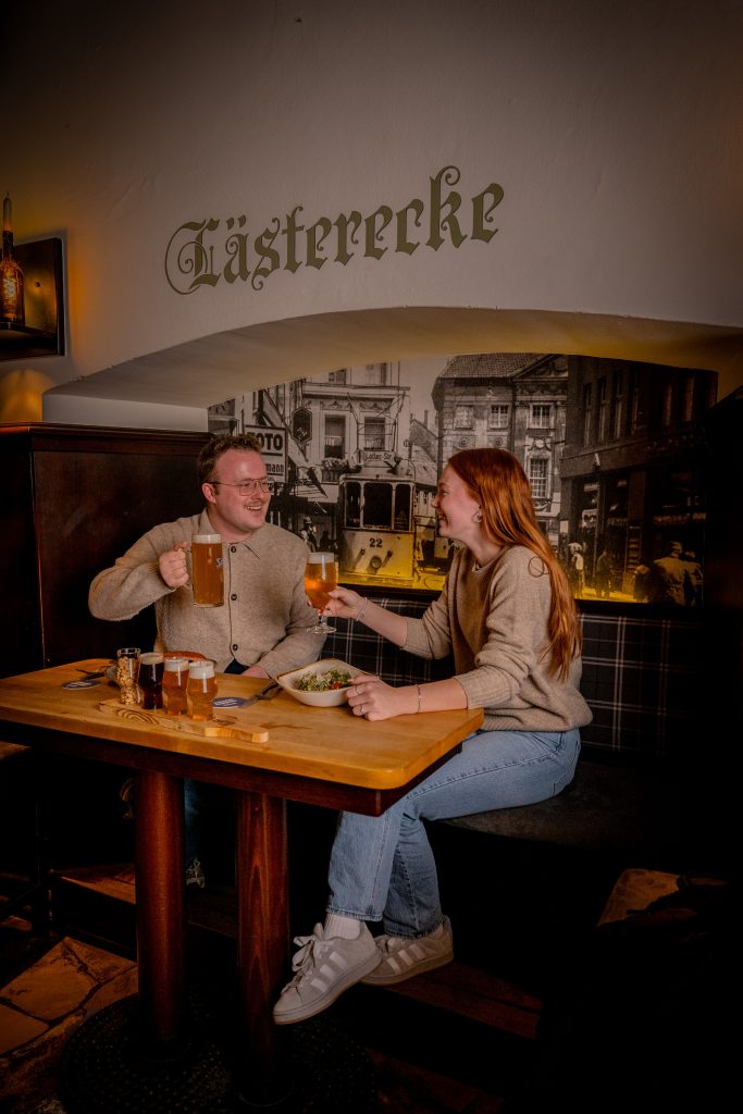 Mann und Frau sitzen in der Lästerecke und prosten sich mit Bier zu